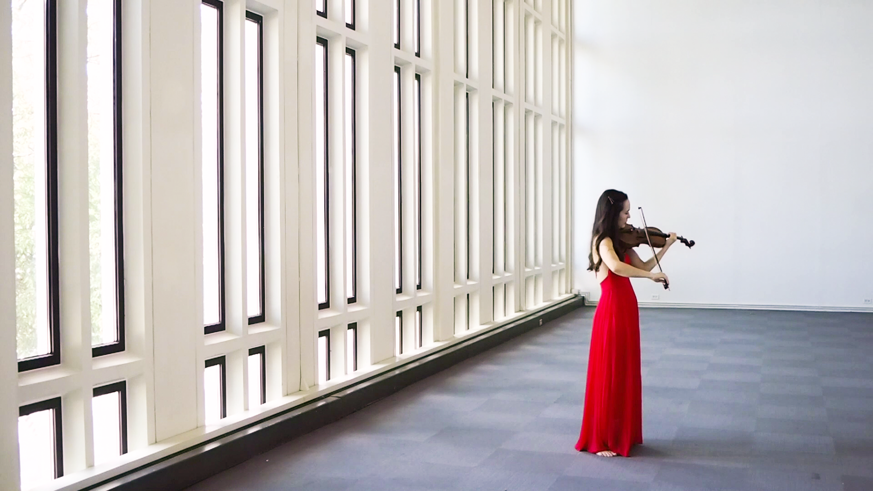 A woman in a red dress playing violin next to a bay of windows