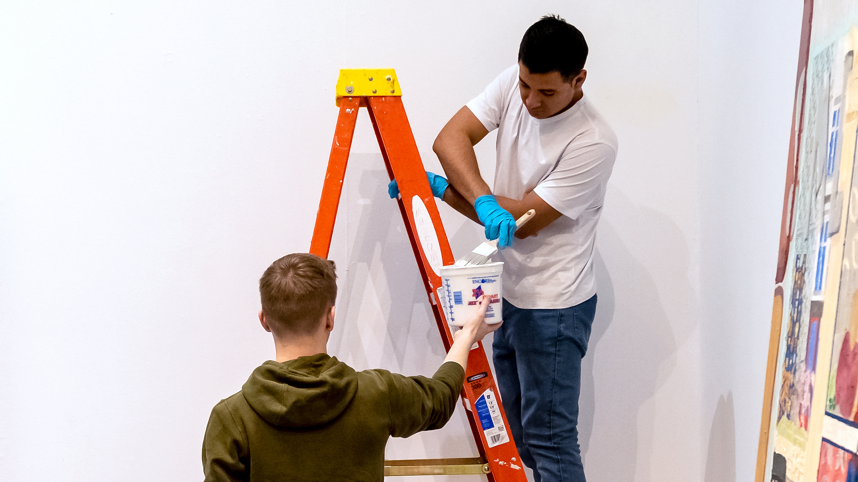 Two college students— one on a ladder with a paint brush and the other holding the paint bucket for his friend