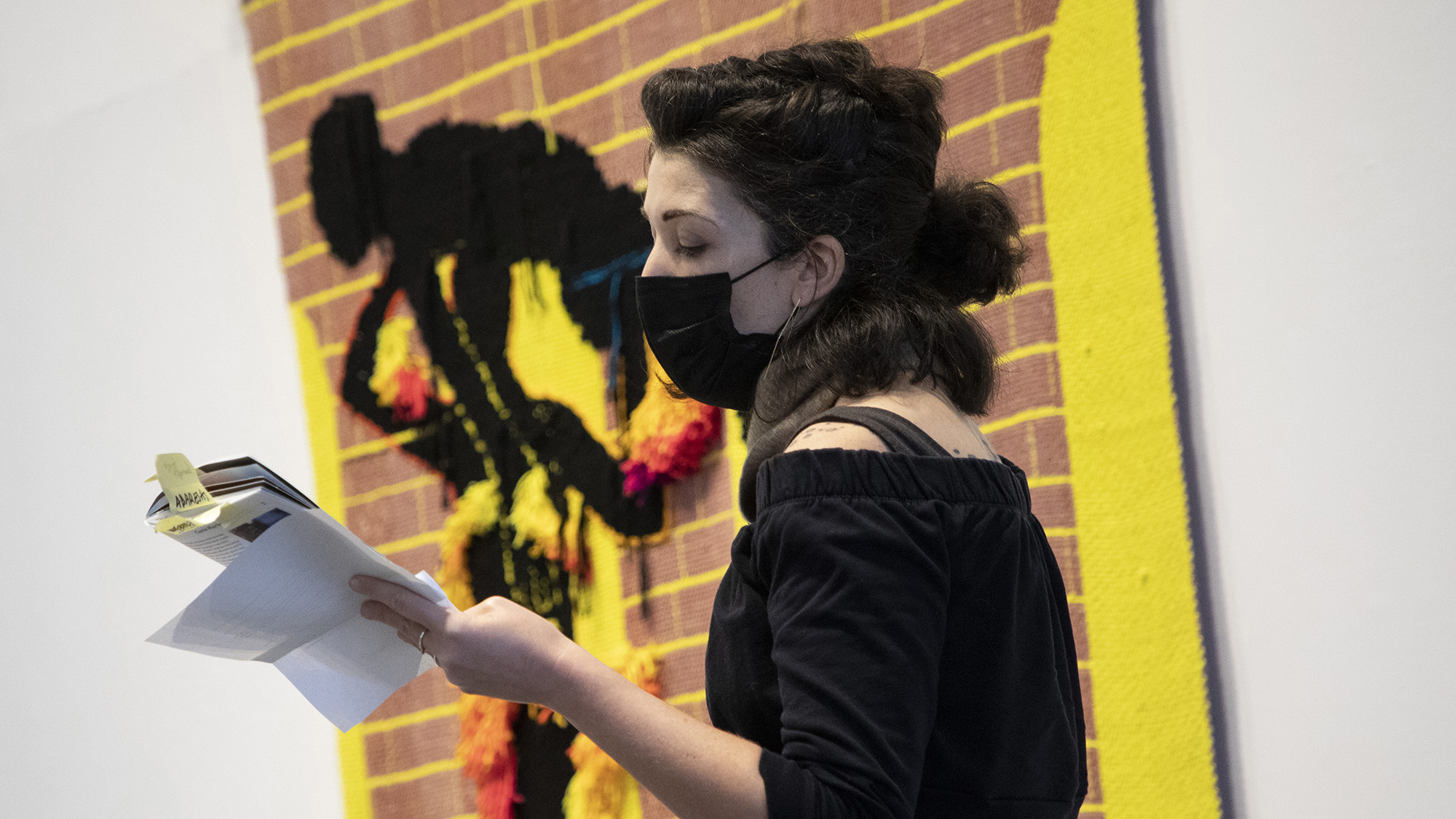 A person in a fabric mask reading from a catalog standing in front of a woven artwork.
