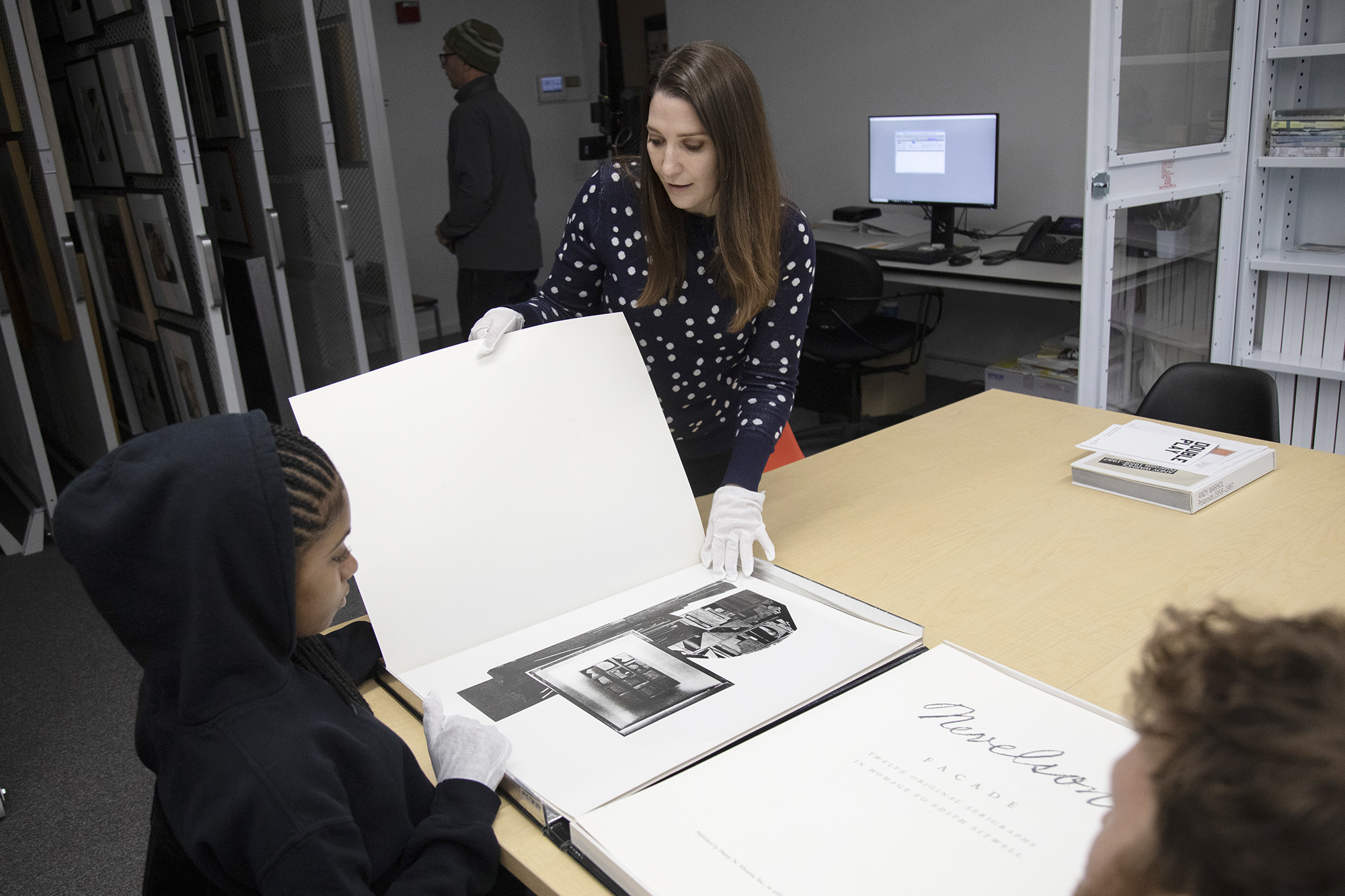 The interior of the Collections Study Space. In the foreground, a young student wearing white gloves and a black hooded sweatshirt is being shown an artist portfolio from a UAM staff member wearing white gloves and a blue sweater with white dots. In the background sits a bank of sliding racks where artwork is attached. 