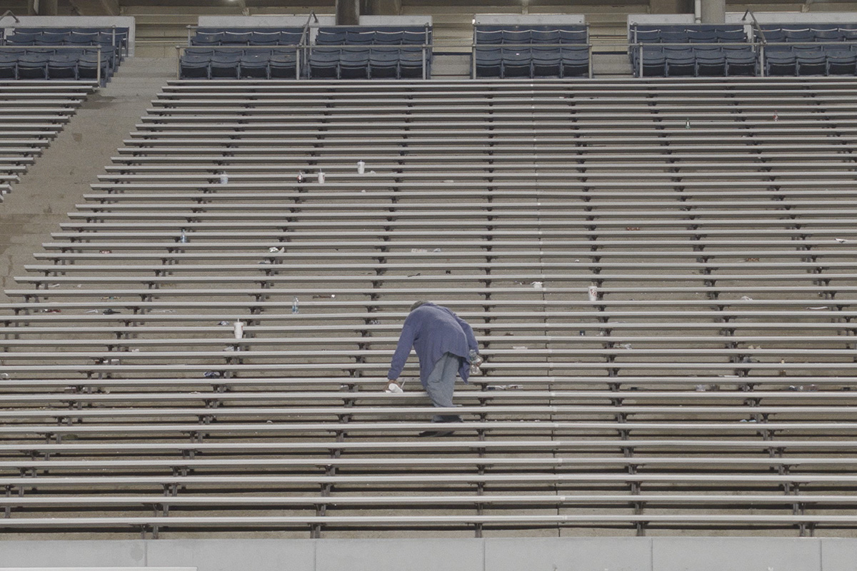 A lone person wearing a gray/blue sweater and gray pants is cleaning debris off a bank of empty stadium bleachers.