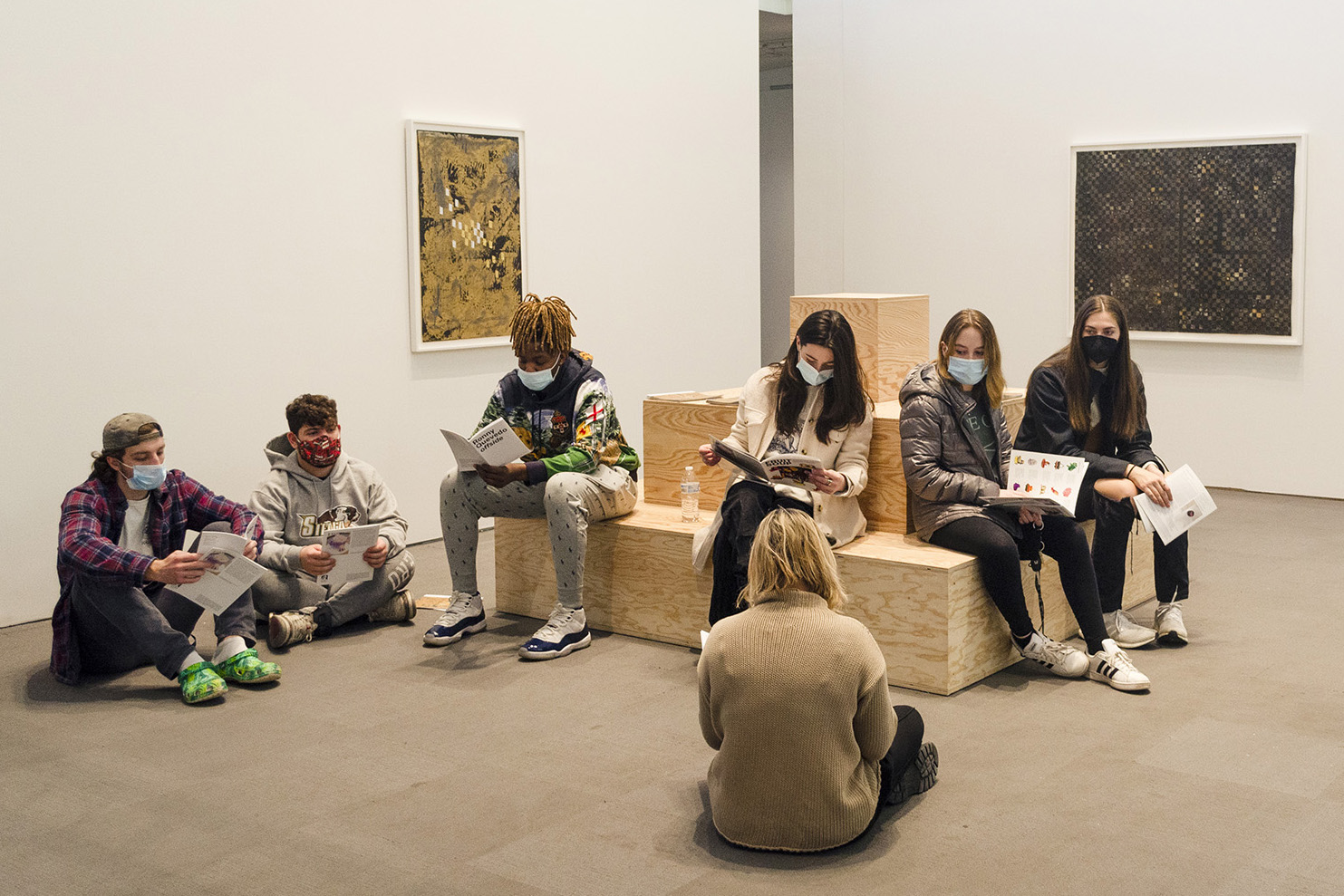 The interior first floor of the University Art Museum. In the foreground a professor wearing a beige sweater sits cross-legged on the ground, their back to us. Six students sit on the ground and on a plywood structure facing the professor. Two  framed artworks are attached to the walls in the background.