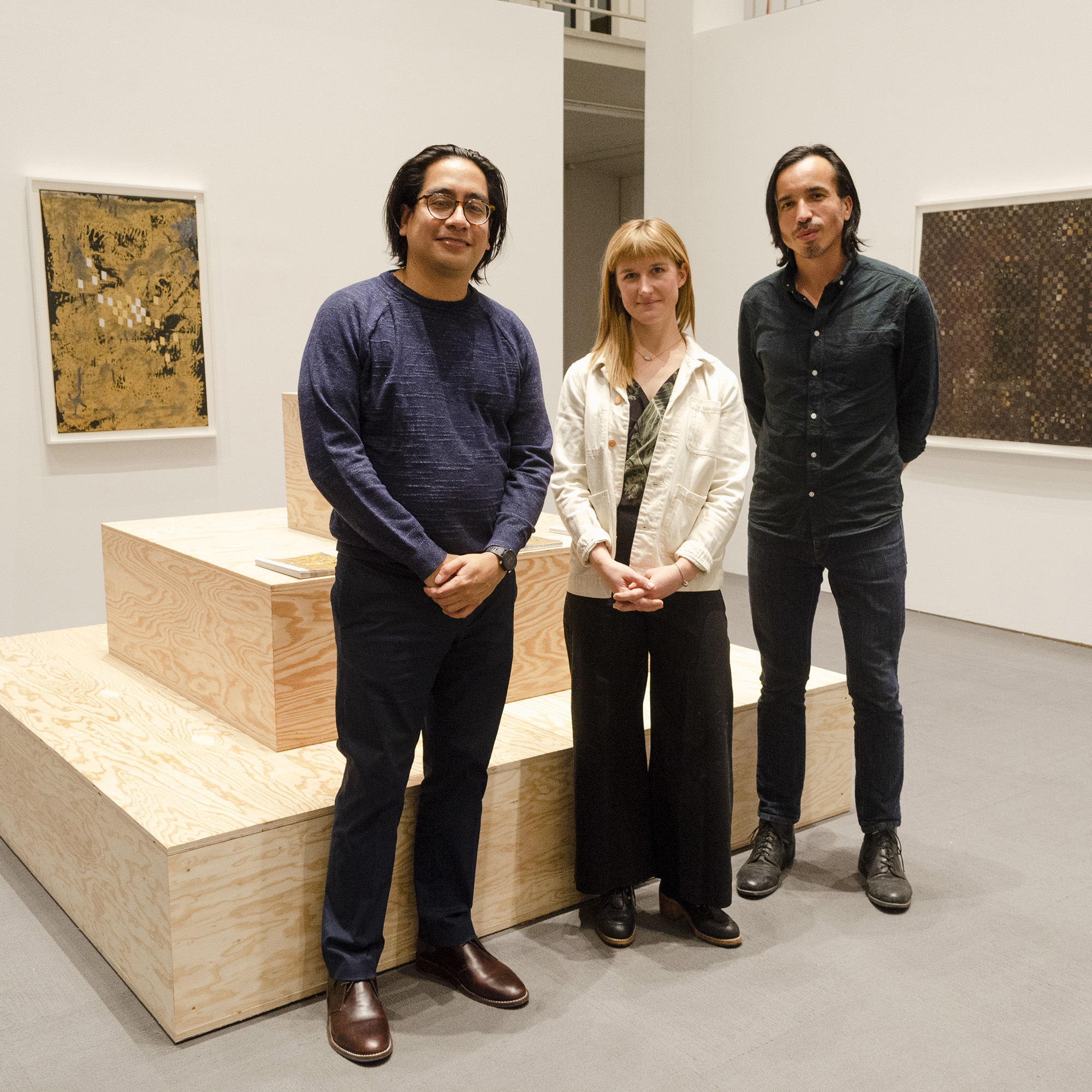 Three artists, Ronny Quevedo, Gracelee Lawrence, and Rodrigo Valenzuela are standing and smiling in front of a large three-tiered plywood sculpture inside the University Art Museum. Two artworks are attached to the wall in the background.